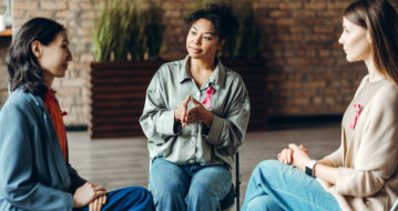 Three people wearing pink ribbons sit in a circle, engaging in a supportive group discussion in a cozy indoor setting.