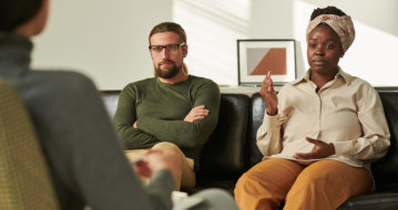 Two people sit on a couch engaged in conversation with a third person, likely in a counseling or therapy session.