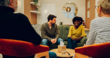 A group of people sitting in a circle in a cozy room, engaged in a discussion. One person appears to be speaking while others listen attentively.