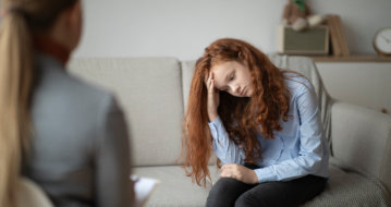 A person with long red hair sits on a sofa, leaning forward with their face resting in their hand, appearing distressed.