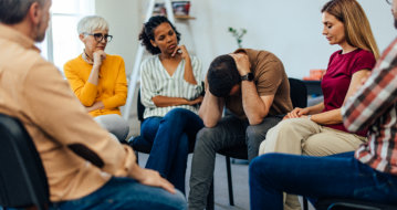 A man sitting with his head in his hands, appearing distressed, while a woman beside him offers comfort by placing her hand on his knee. Other people are seated nearby.