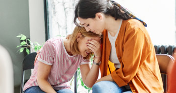 Two women sit close indoors, one comforting the other who appears upset, holding her face in her hand.