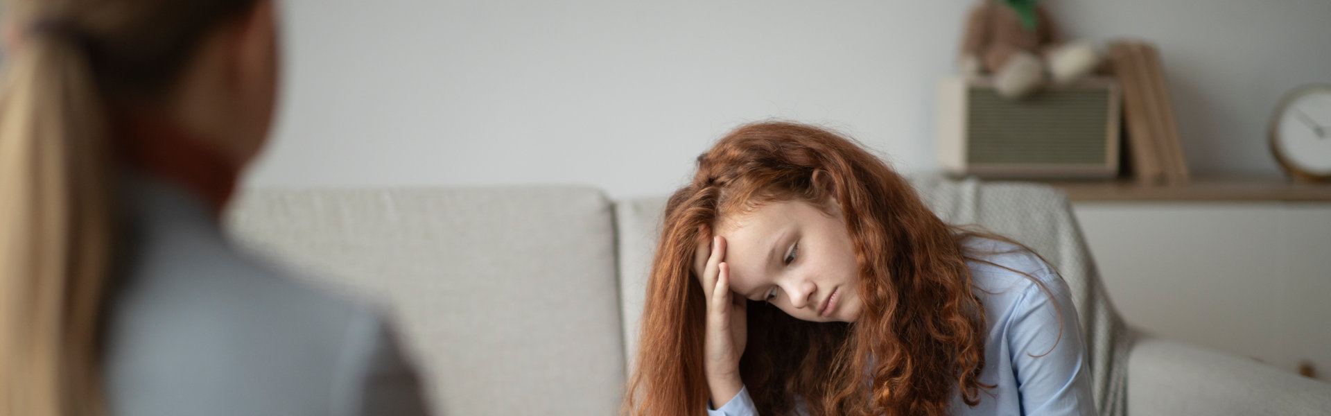 A person with long, curly hair sits on a couch, holding their head in their hand, appearing distressed. Another person is seated across from them.