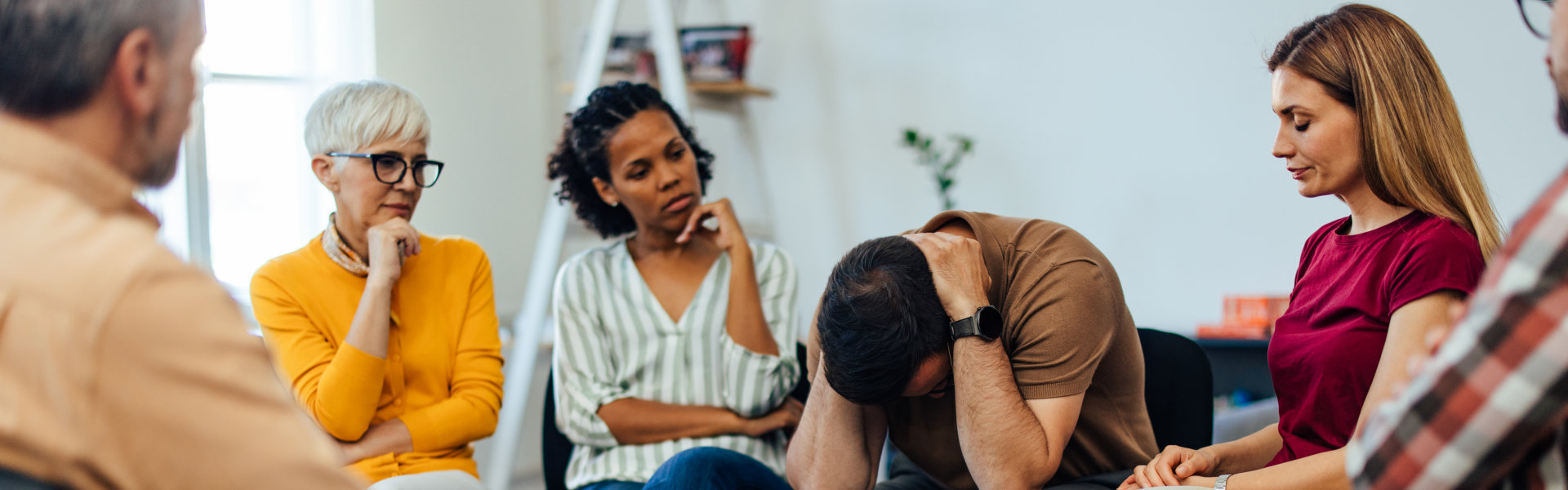 A group of people sitting in a circle, engaged in a discussion. One person appears distressed, with their head in their hands.